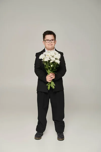 Boy with down syndrome in school uniform and eyeglasses holding flowers and smiling on grey — Stock Photo