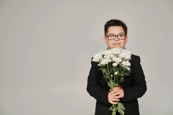 Happy schoolboy in uniform and eyeglasses standing with flowers on grey, inclusive schooling — Stock Photo