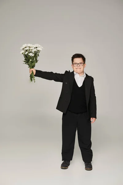Schoolboy with down syndrome in eyeglasses and uniform holding bouquet in outstretched hand on grey — Stock Photo