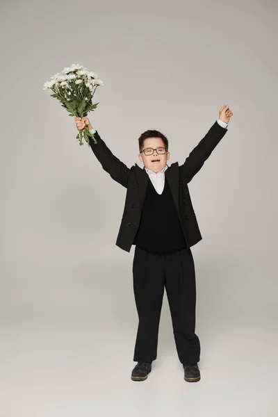 Взволнованный школьник с синдромом Дауна, стоящий с цветами и поднятыми руками на сером, полной длины — стоковое фото