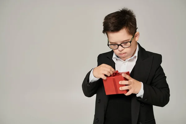 Мальчик с синдромом Дауна в школьной форме и очках открывает подарочную коробку с красной лентой на сером — стоковое фото