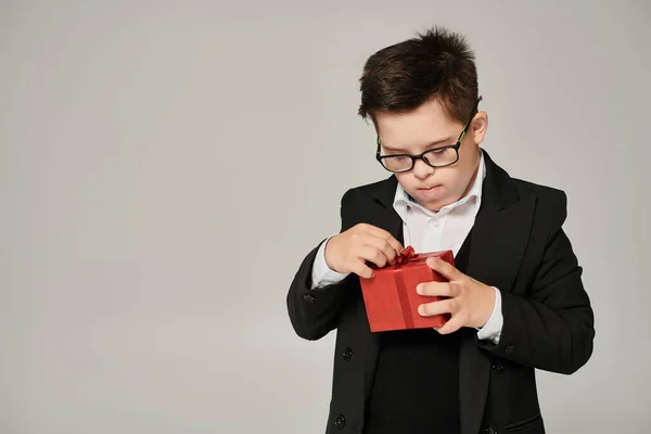 Junge mit Down-Syndrom in Schuluniform und Brillenöffnung Geschenkbox mit roter Schleife auf grau — Stockfoto