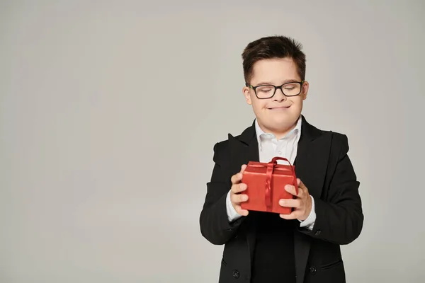 Довольный школьник с умственной отсталостью держит красную подарочную коробку на сером, синдром Дауна концепции — стоковое фото