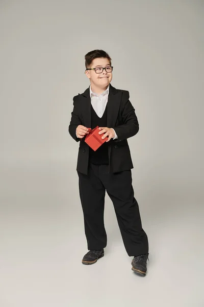 Веселий хлопчик з синдромом Дауна в шкільній формі і окуляри стоять з червоною подарунковою коробкою на сірому — стокове фото