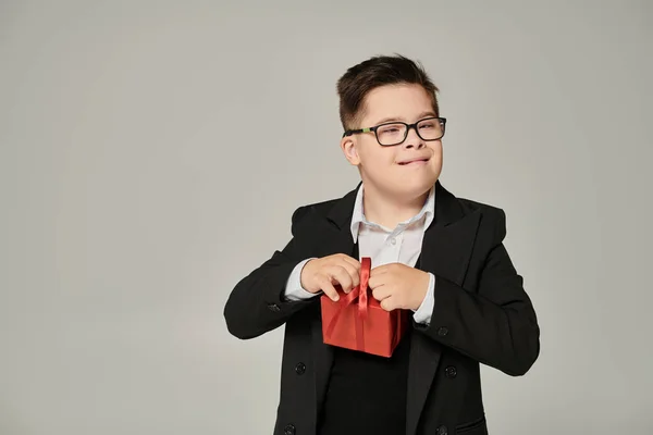 Kind mit Down-Syndrom in Brille und Schuluniform mit Geschenkverpackung auf grau, Sonderschüler — Stockfoto