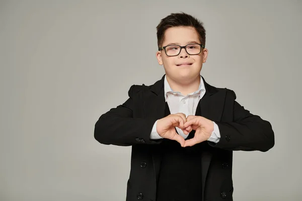 Niño feliz con síndrome de Down en uniforme escolar y gafas que muestran signo de amor con las manos en gris - foto de stock
