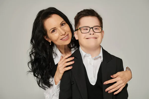 Donna di mezza età che abbraccia il figlio con la sindrome di Down in uniforme scolastica mentre sorride alla telecamera sul grigio — Foto stock