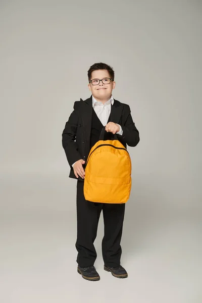 Cheerful schoolboy with down syndrome, in black suit and eyeglasses holding yellow backpack on grey — Stock Photo