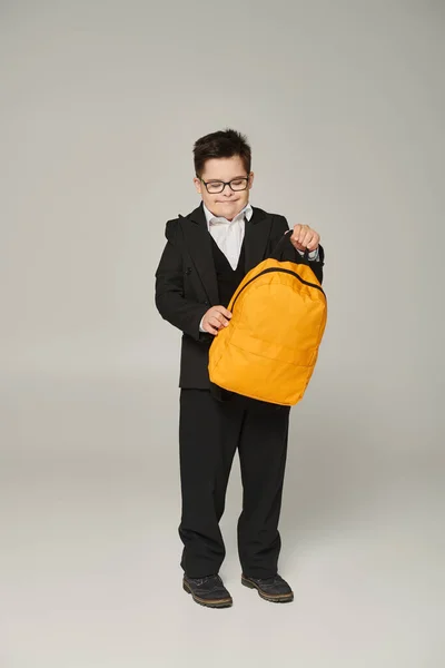 Happy schoolboy with down syndrome, in black uniform and eyeglasses holding yellow backpack on grey — Stock Photo