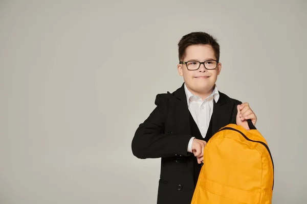 Школярка з синдромом Дауна тримає жовтий рюкзак і посміхається на сірому, інклюзивному шкільному — стокове фото