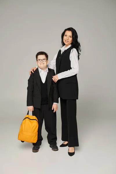 Щасливий школяр з синдромом Дауна, з жовтим рюкзаком поруч і матір'ю в офіційному одязі на сірому — стокове фото