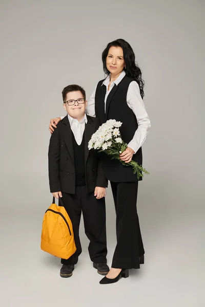 Жінка з квітами і школярка з синдромом вниз тримає жовтий рюкзак на сірій, спеціальній сім'ї — стокове фото