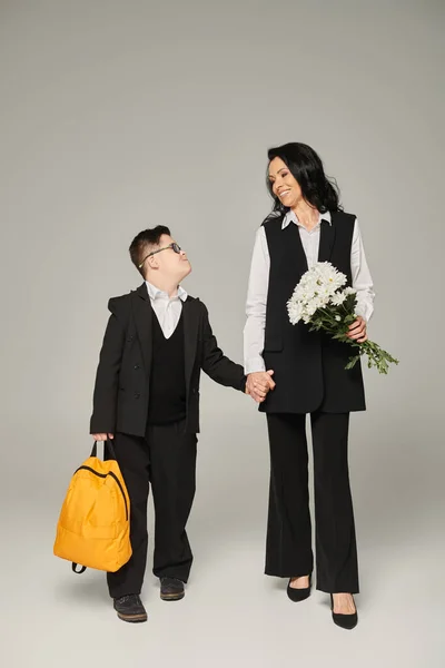 Schoolboy with down syndrome holding yellow backpack near mother with flowers on grey, full length — Stock Photo