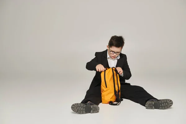 Школьник с синдромом Дауна, в черном костюме и очках сидит с желтым рюкзаком на сером — стоковое фото