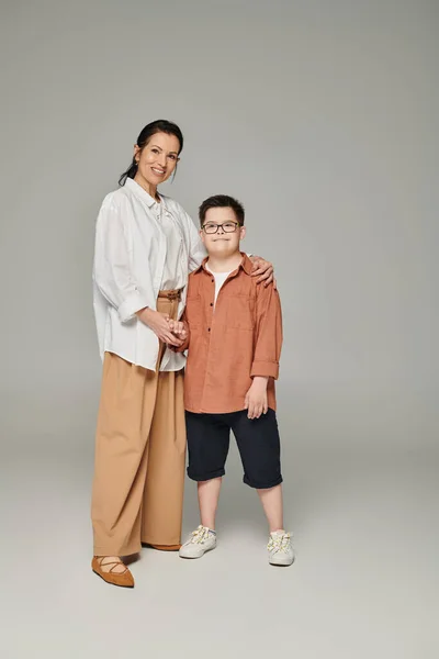 Happy boy with down syndrome, in eyeglasses, holding hands with mother smiling at camera on grey — Stock Photo