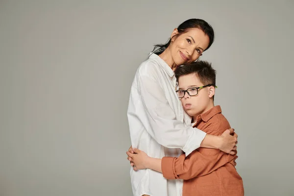 Joyful middle aged woman hugging son with down syndrome and smiling at camera on grey, banner — Stock Photo