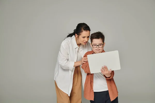 Mujer de mediana edad explicando lección en línea para sonreír hijo con síndrome de Down cerca de la computadora portátil en gris - foto de stock