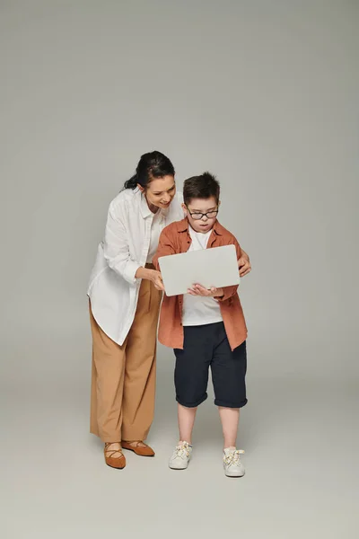 Sorridente donna di mezza età che spiega lezione online al figlio con sindrome di Down vicino laptop su grigio — Foto stock