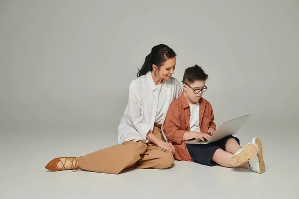 Kid with down syndrome sitting near smiling mother and using laptop on grey, full length — Stock Photo
