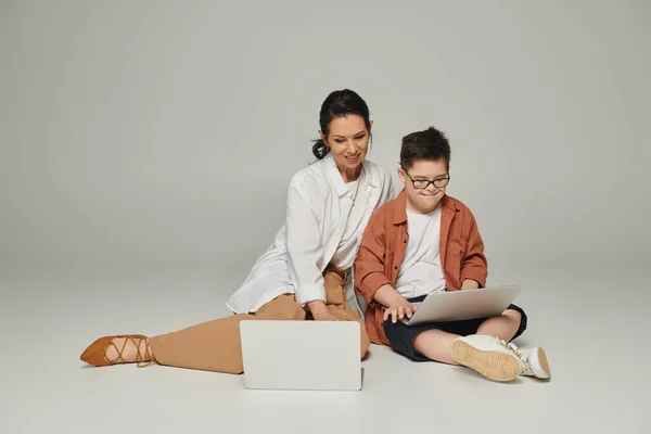 Positive middle aged woman and boy with down syndrome sitting with laptops on grey, special family — Stock Photo