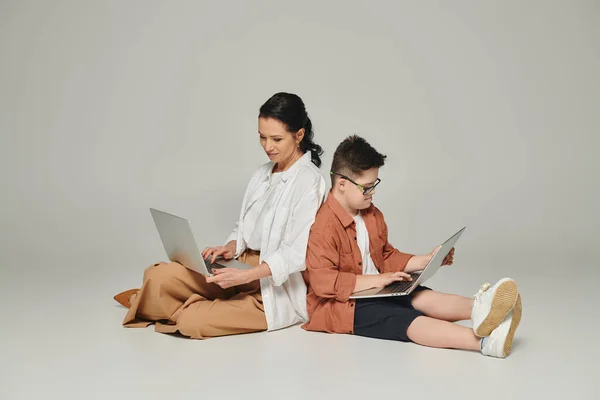 Femme d'âge moyen et enfant avec le syndrome du duvet assis dos à dos avec des ordinateurs portables sur gris — Photo de stock