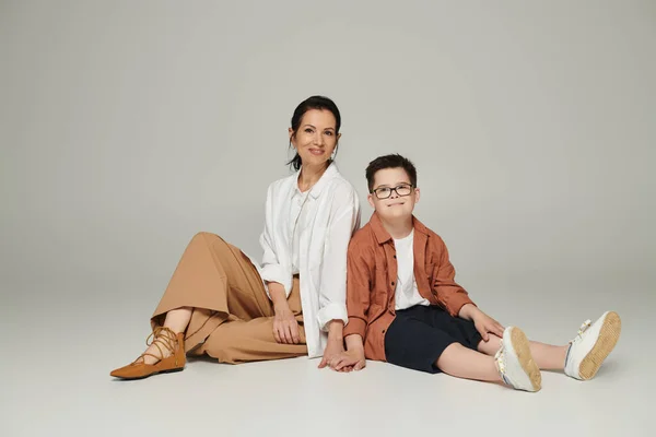 Menino alegre com síndrome de down e mãe de meia-idade em roupas elegantes sorrindo e sentado em cinza — Fotografia de Stock