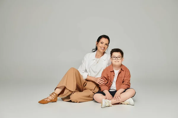 Middle aged woman and boy with down syndrome sitting and looking at camera on grey, special family — Stock Photo