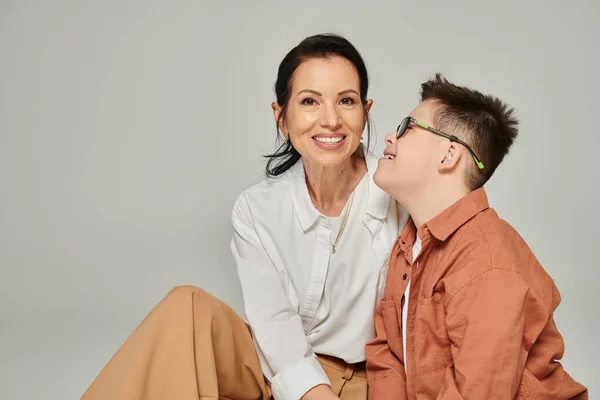 Жінка середнього віку посміхається біля радісного сина з синдромом Дауна на сірому, прийняття інвалідності — стокове фото