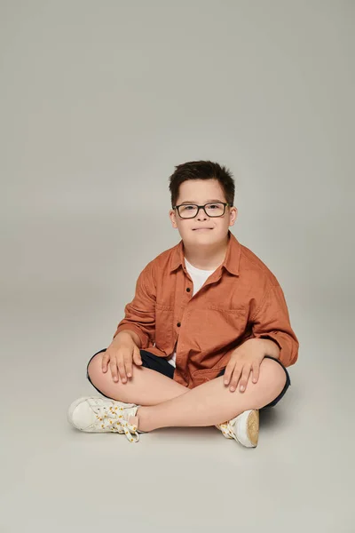 Niño feliz con síndrome de Down en ropa casual de moda y gafas de vista sentado y sonriendo en gris - foto de stock