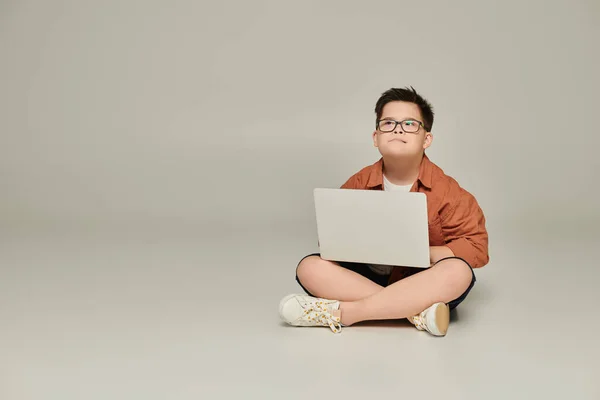 Стильный и вдумчивый мальчик с синдромом Дауна сидит с ноутбуком и скрещенными ногами на сером — стоковое фото