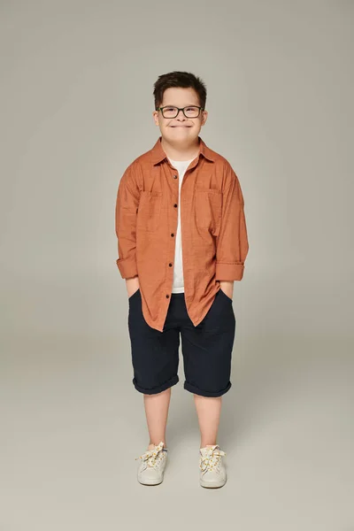 Niño alegre con síndrome de Down en pantalones cortos y anteojos posando con las manos en bolsillos en gris - foto de stock