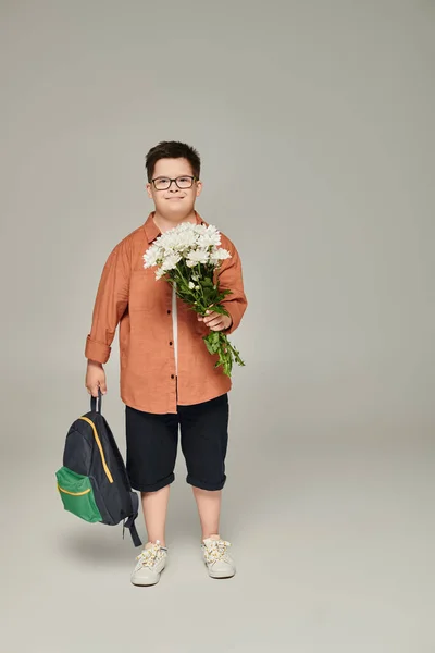 Joyful boy with down syndrome, in casual attire and eyeglasses holding backpack and flowers on grey — Stock Photo