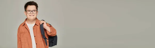 Schoolboy with down syndrome in stylish clothes and eyeglasses holding backpack on grey, banner — Stock Photo