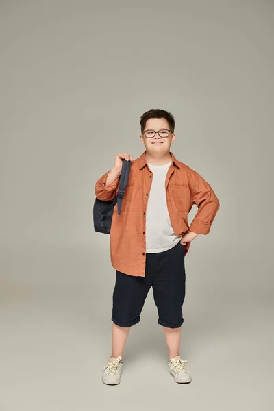 Menino sorridente com síndrome de down, em roupas da moda, posando com mochila escolar em cinza — Fotografia de Stock