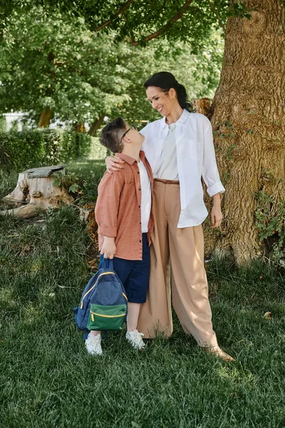 Щаслива жінка середнього віку, що приймає сина з синдромом Дауна, тримає рюкзак в парку, повна довжина — стокове фото