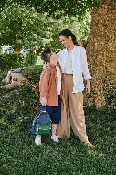 Happy middle aged woman embracing son with down syndrome holding backpack in park, full length — Stock Photo