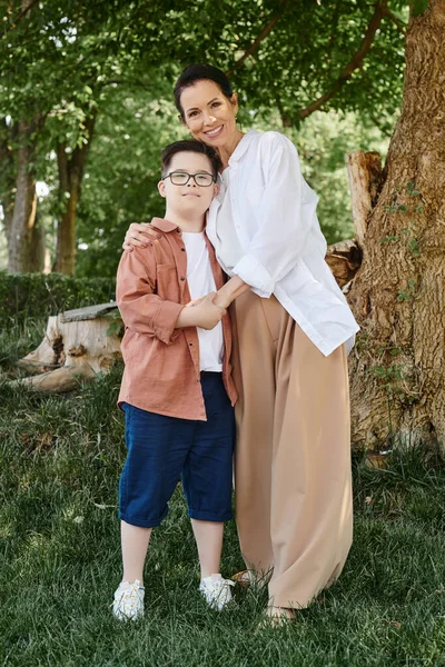 Joyful middle aged woman and son with down syndrome hugging and holding hands in green park — Stock Photo