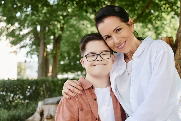 Middle aged woman smiling at camera and hugging pleased son with down syndrome in park — Stock Photo