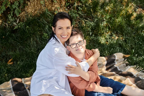 Felice donna di mezza età seduta su una coperta nel parco e abbracciare il figlio con la sindrome di Down, tempo di qualità — Foto stock