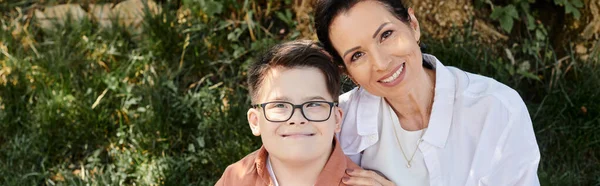 Unbekümmerter Junge mit Down-Syndrom lächelt neben glücklicher Mutter mittleren Alters im grünen Park, Banner — Stockfoto
