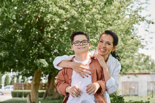 Веселая женщина средних лет обнимает улыбающегося сына с синдромом Дауна в парке, безусловная любовь — стоковое фото