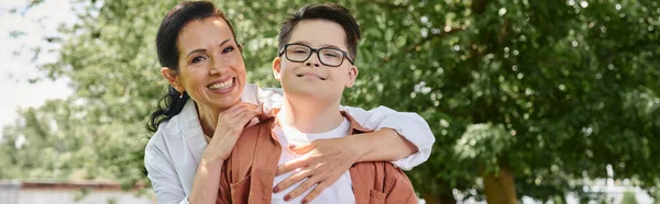Sonriente mujer de mediana edad abrazando hijo con síndrome de Down en el parque, conexión emocional, pancarta - foto de stock