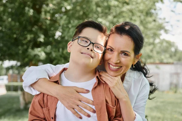 Pleased middle aged woman embracing boy with down syndrome and smiling in park, special family — Stock Photo