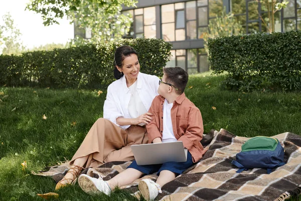 Smiling woman and boy with down syndrome sitting near laptop on blanket in park, unique family — Stock Photo