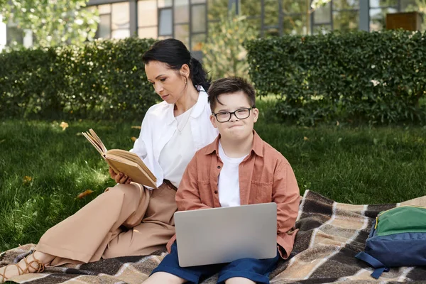 Femme d'âge moyen lecture livre près de fils avec le syndrome du duvet et ordinateur portable sur couverture dans le parc, loisirs — Photo de stock