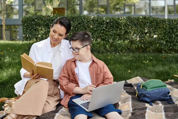 Happy middle aged woman reading book near son with down syndrome using laptop in park — Stock Photo