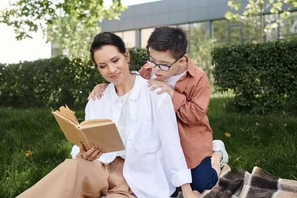 Усміхнена жінка середнього віку читає книгу синові з синдромом Дауна в парку, якісний час, дозвілля — стокове фото
