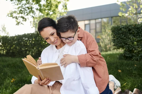 Mulher de meia-idade alegre lendo livro para filho com síndrome de down durante o lazer no parque — Fotografia de Stock
