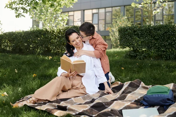 Junge mit Down-Syndrom küsst glückliche Mutter beim Lesen von Buch auf Decke im Park, emotionale Verbindung — Stockfoto