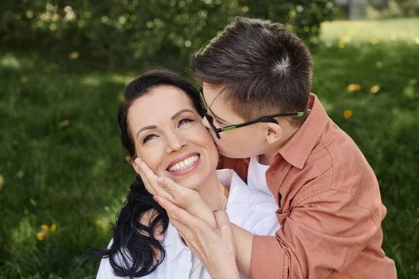Мальчик-подросток с синдромом Дауна, в очках, целующийся с довольной матерью в парке, безусловная любовь — стоковое фото